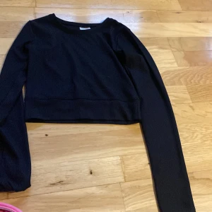 Svart croppad tröja från Kappahl - Svart croppad långärmad tröja från Kappahl. Tröjan har en ribbad struktur och rund halsringning, perfekt för en chill och stilren look. Passar dig som gillar enkla basplagg med en modern twist.