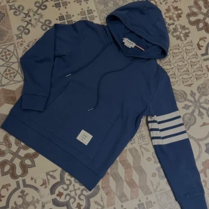 Rare hoodie från Thom Browne (Rea idag) - Snygg blå hoodie från Thom Browne med vita ränder på ena ärmen och klassisk huva med snörning. Tröjan har en känguruficka framtill och en vit etikett nertill. Perfekt för dig som gillar stilrena och sportiga plagg. Tröjan är i storlek 3 vilket motsvarar M. Priset utgår ifrån vad den går för på andra marknader! Kan gå ned i pris vid blixtsnabb affär!
