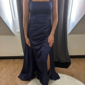 Marinblå långklänning med slits - Elegant marinblå långklänning i glansigt material med breda axelband och fyrkantig urringning. Klänningen har en snygg slits framtill och en figurnära överdel som övergår i ett svepande kjolparti. Perfekt för bal eller festliga tillfällen.