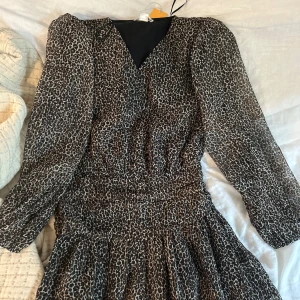 Leopardmönstrad kortklänning med glitter - Supersnygg kort klänning från Gina med leopardmönster och diskreta glittriga trådar. Klänningen har v-ringning, långa ärmar med lätt puff och smockad midja som ger en schysst siluett. Perfekt för dig som vill sticka ut lite extra.