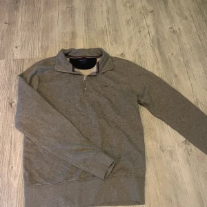 Grå half zip tröja från GANT - Snygg grå tröja från GANT med half zip. Tröjan har diskret broderad logga på bröstet och randiga detaljer i kragen. Perfekt för lager-på-lager och chill stil. Tillverkad i mjuk bomull för skön känsla.