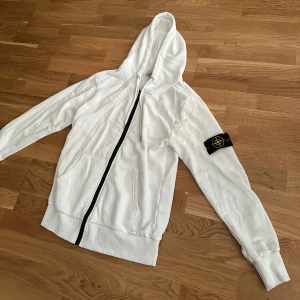 Vit hoodie från Stone Island med dragkedja - Vit hoodie från Stone Island med svart dragkedja, huva med snörning och klassisk logotyp-patch på vänster ärm. Tröjan har känguruficka framtill och är tillverkad i mjuk bomull. Perfekt för dig som gillar clean och stilren streetwear.