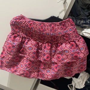 Rosa mönstrad kortkjol med volang - Supersöt kortkjol i rosa med färgglatt mönster och bred smock i midjan. Kjolen har volang och är perfekt för sommarens alla äventyr. Lätt och luftig känsla, passar dig som gillar färg och mönster. Och har undershorts. Kjolen är i storlek XS men den passar som i storlek S/M