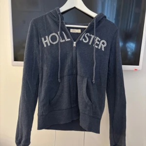 Mörkblå hoodie från Hollister - Mörkblå hoodie från Hollister med vit broderad logga över bröstet. Tröjan har huva med snörning, dragkedja framtill och två fickor💓