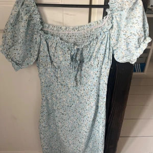 Blommig ljusblå klänning - Jättesöt ljusblå klänning med blommigt mönster och puffiga kortärmar. Den har en smockad överdel och en söt knytning framtill. Den sitter åt vid midjan och höfterna och går ner till mitten på låren. Supersomrig🩵🤍Osäker på storlek men den passar både för S och XS! 