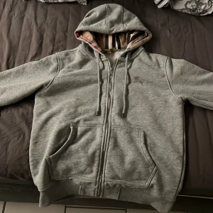 Burberry hoodie - Säljer en grå hoodie med dragkedja och broderad logga på bröstet. Den har en justerbar huva med snören och praktiska fickor framtill. Perfekt för en avslappnad stil. Pris kan diskuteras
