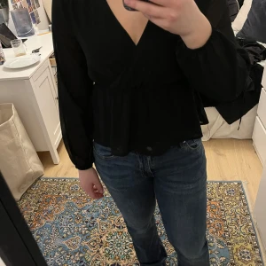 Svart omlottblus från Vila - Snygg svart omlottblus från Vila med långa ärmar och spetsdetaljer på axlarna. Blusen har en v-ringad design och elastisk midja för en smickrande passform. Perfekt för en stilren look.