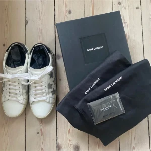 Vita sneakers med stjärnor från Saint Laurent - Snygga vita sneakers från Saint Laurent med silverfärgade stjärnor på sidorna. Skorna har en klassisk snörning och en bekväm sula. Perfekta för att ge din outfit en cool touch. Tyvärr tillkommer inga sulor, fick inte med några.