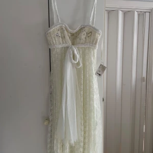 Vit Balklänning - Elegant vit långklänning i spets med rätt tjocka axelband och dekorativa paljetter. Klänningen har en knytning framtill och en vacker, genomskinlig spetsöverdel. Perfekt för speciella tillfällen. Prislappen är även kvar så den är ej använd - Originalpriset: 2800kr