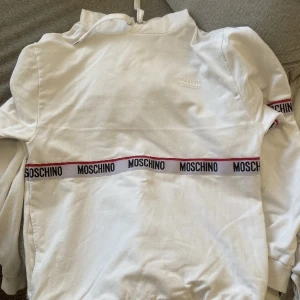 Vit hoodie från Moschino - Säljer en stilren vit hoodie från Moschino med deras logga tryckt på bröstet och längs ärmarna. Hoodien har en klassisk design med dragsko i huvan och ribbade muddar. Perfekt för en avslappnad look. Kvitto till hoodien finns och den ser helt ny ut