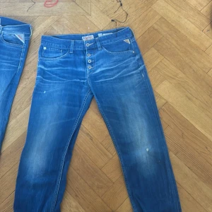 Blå jeansbyxor från Replay - Snygga blå jeansbyxor från Replay med knappar framtill och klassisk femficksdesign. De har en lätt slitning för en cool look och märkets logga på bakfickan. Perfekta för en avslappnad stil.