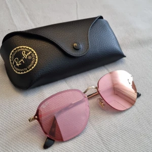 Solglasögon med rosa linser från Ray-Ban - Solglasögon från Ray-Ban (äkta) med rosa linser och guldfärgade bågar. De har en unik geometrisk form som ger en modern look. Kommer med ett stilrent svart fodral för säker förvaring. Aldrig använda.  