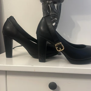 Svarta pumps med klack 35 - Snygga svarta pumps med klack och en elegant rem med guldfärgat spänne. Perfekta för en stilren look. Tillverkade i ett slätt material som ger en klassisk känsla.