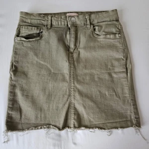 Khaki jeanskjol från Kids Only - Snygg olivgrön jeanskjol från Kids Only med rå kant och klassisk femficksdesign. Kjolen har en dragkedja och knapp framtill för enkel påklädning. Perfekt för en avslappnad stil. Bara anvånd npgra gånger. Bara att fråga om du har någon fundering💗