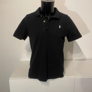 Svart pikétröja från Ralph Lauren - Snygg svart pikétröja från Ralph Lauren med klassisk krage och knappslå. Tröjan har en broderad logotyp på bröstet och korta ärmar. Perfekt för en stilren look.