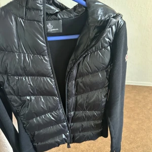 Svart jacka från Moncler - Snygg svart jacka från Moncler med glansig pufferdesign framtill och mjuka ärmar. Jackan har en dragkedja och en hög krage för extra stil och värme. Perfekt för kyligare dagar när du vill hålla stilen.
