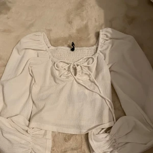 Vit blus med knytning - Säljer en söt vit blus med långa puffiga ärmar och knytning framtill. Blusen har en smockad design på baksidan och är croppad för en trendig look.