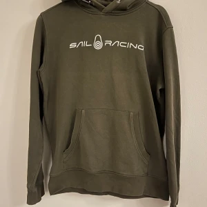 Olivgrön hoodie från Sail Racing - Säljer en olivgrön hoodie från Sail Racing med vit logotyp på bröstet. Tröjan har en klassisk känguruficka och justerbar huva med dragsko. Perfekt för en avslappnad stil.