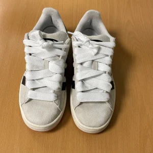 Vita Adidas Campus sneakers - Snygga vita Adidas Campus sneakers med svarta detaljer och klassiska tre ränder på sidan. Skorna har breda vita snören och en bekväm passform. Perfekta för en stilren look.