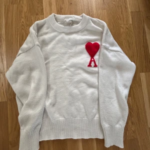 Vit ami pris  tröja med rött hjärta - Säljer en vit tröja med ett rött hjärta och bokstaven A på bröstet. Tröjan har långa ärmar och ribbade kanter vid ärmslut och nederkant. Perfekt för en stilren look! Köpt från CN fans 