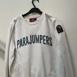 Vit tröja från Parajumpers - Säljer en vit tröja från Parajumpers med tryckt logotyp på framsidan och ett märke på ärmen. Tröjan har långa ärmar och dekorativa sömmar i orange vid axlarna. Perfekt för en stilren och avslappnad look. Passar S/M ✅ Pris:300kr