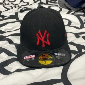 Svart keps från New Era - Snygg svart keps från New Era med röd broderad logga framtill. Kepsen är i modellen 59FIFTY och har en fast storlek på 7 (55.8 cm). Perfekt för att ge din outfit en sportig touch.