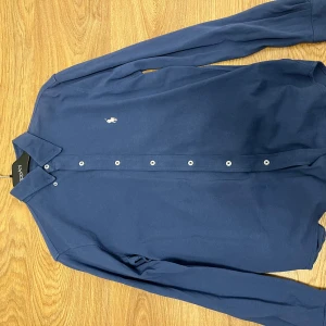 Ralph Laurent skjorta - Snygg blå långärmad skjorta från Ralph Lauren med klassisk krage och knappar framtill. Skjortan har en broderad logotyp i rosa på bröstet. Perfekt för en stilren looken.