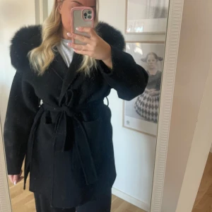 Svart kappa med pälsdetaljer - Säljer en elegant svart kappa med lyxig pälskrage och knytbälte i midjan. Perfekt för att hålla sig varm och stilren under kyliga dagar. Den har långa ärmar och en klassisk design som passar till många tillfällen. Från femeninaofsweden, ord pris 3799kr säljer för 1800kr