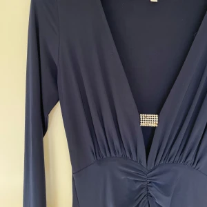 Långklänning, bal/nyår/fest med djup v-ringning och slits - Elegant mörkblå långklänning med djup v-ringning och dekorativt spänne strax ovanför midjan för att hålla ihop v-ringningen. Klänningen har långa ärmar och ett stilrent, draperat fall framtill. Hög slits. Smickrande passform iom scrunchy framsida. Använd endast vid ett tillfälle.