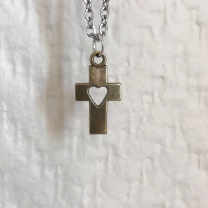 Halsband med kors och hjärta - Snyggt halsband med ett korshänge i metall. Hänget har ett utskuret hjärta i mitten och hänger på en silverfärgad kedja. Perfekt för att ge en stilren touch till din outfit.