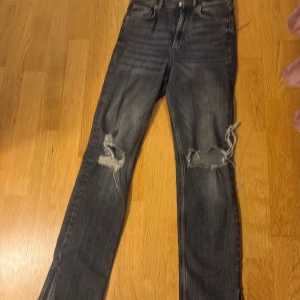 Gråa jeans med slitningar från Zara - Snygga gråa jeans från Zara med slitningar på knäna och slits på sidorna. De har en klassisk femficksdesign och knappgylf. 