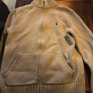 Grå stickad kofta från Ralph Lauren storlek 14/16 - Säljer en stilren beige stickad kofta från Ralph Lauren med dragkedja och ribbad krage. Den har långa ärmar och praktiska fickor framtill. Perfekt för kyligare dagar!