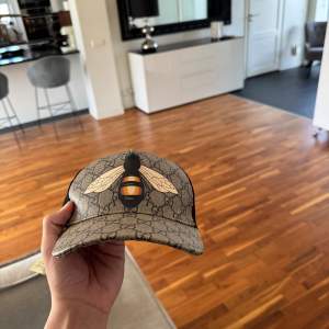 Snygg beige keps från Gucci med ett stort bi-motiv på framsidan. Kepsen har ett klassiskt mönster och justerbart band baktill i grönt och rött. Perfekt för att ge din outfit en unik touch.
