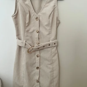 Beige ärmlös klänning med bälte - Snygg beige ärmlös klänning med knappar framtill och ett matchande bälte i midjan. Klänningen har en v-ringad design och ett subtilt randigt mönster. Perfekt för en stilren look!