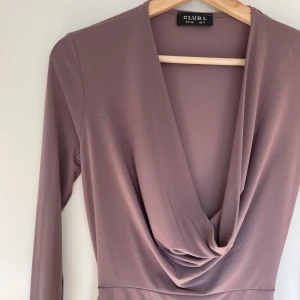 Mörkrosa klänning från Club L - Snygg mörkrosa klänning från Club L med draperad v-ringning och lång ärm. Perfekt för en elegant look. Klänningen har en figurnära passform som framhäver silhuetten.