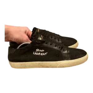 Svarta sneakers från Saint Laurent - Snygga svarta sneakers från Saint Laurent med vit logga på sidan. Skorna har en klassisk design med snörning och en bekväm sula. Perfekta för en stilren look. Priset är inte hugget i sten kan sänkas till 1700kr om det behövs. Byten går att göras! 