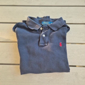 Blå pikétröja från Ralph Lauren - Säljer en klassisk blå pikétröja från Ralph Lauren med lång ärm och den ikoniska röda broderade loggan på bröstet. Perfekt för en stilren och avslappnad look. Tröjan har knappar vid kragen och är i ett mjukt bomullsmaterial.