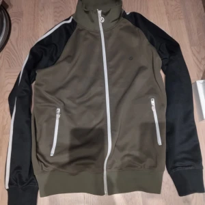 Olivgrön jacka från Sol Jeans - Säljer en olivgrön jacka från Sol Jeans med svarta ärmar och vita ränder. Jackan har dragkedja framtill och två fickor med dragkedjor. Perfekt för en sportig look!