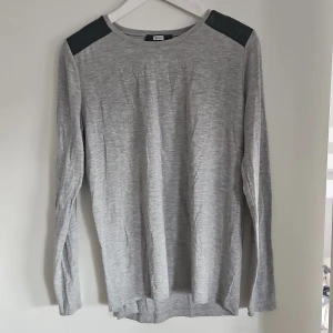 Grå långärmad tröja med quiltad detalj - Snygg grå långärmad tröja med svarta quiltade detaljer på axlarna och övre ryggen. Perfekt för en stilren look med en twist. Passar bra till både jeans och kjol.