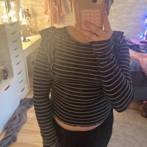 Svart och vit randig topp - Säljer en svart och vit randig topp med långa ärmar och volangdetaljer vid axlarna. Toppen har en croppad passform och är perfekt för en trendig look. Hör av dig vid intresse!