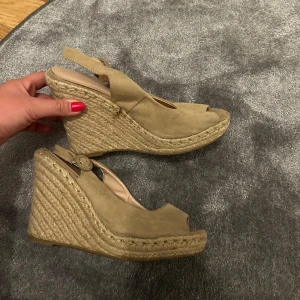 Beige kilklackssandaler - Snygga beige sandaler med kilklack och öppen tå. De har en flätad detalj runt sulan och en justerbar rem runt hälen. Storlek 38