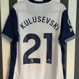 Vit och blå fotbollströja - Säljer en vit och blå fotbollströja med korta ärmar. Tröjan har ett tryck med namnet 'Kulusevski' och nummer 21 på ryggen. Perfekt för fotbollsfans som vill visa sitt stöd.