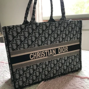 Mönstrad shoppingväska - Snygg shoppingväska med ett ikoniskt mönster i svart och grått. Väskan har två handtag och en stilren design med märkets namn i mitten. Perfekt för att bära med sig allt man behöver med stil.