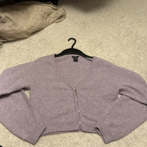 Lila kofta från Lindex - Säljer en söt lila kofta från Lindex i storlek XS. Koftan är croppad med en v-ringning och knappar framtill. Perfekt för lager-på-lager och att hålla sig varm med stil.