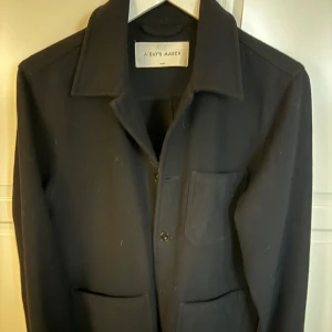Svart overshirt från A Day's March - Snygg svart overshirt från A Day's March med knappar framtill och praktiska fickor. Perfekt för lager-på-lager-stil. Klassisk design med krage och långa ärmar.Storlek S