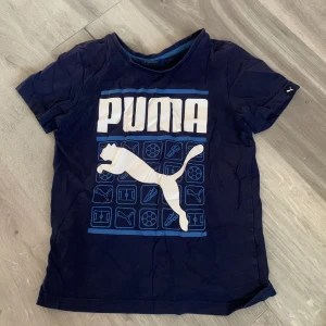 Mörkblå t-shirt från Puma - Säljer en mörkblå t-shirt från Puma med stort vitt tryck och logga på framsidan. Perfekt för sportiga aktiviteter eller vardagsbruk. T-shirten har korta ärmar och är tillverkad i ett bekvämt material. *Barn size 