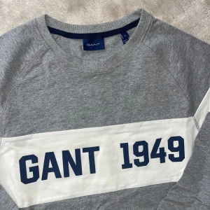 Grå sweatshirt från GANT - Snygg grå sweatshirt från GANT med vit panel och blå text 'GANT 1949' på bröstet. Tröjan har långa ärmar och rund halsringning, perfekt för en avslappnad stil. Finns i st S och L . Kom med bud! 
