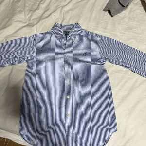 Blå och vit randig skjorta från Ralph Lauren - Snygg blå och vit randig skjorta från Ralph Lauren. Skjortan har långa ärmar och knappar framtill. Perfekt för en stilren look. Passar xxs/xs