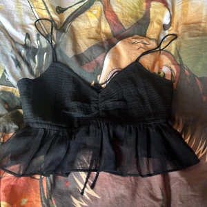 Svart topp från Gina Tricot - Snygg svart topp från Gina Tricot med smala axelband och volangdetaljer. Toppen har en ribbad struktur och knytning i ryggen för en justerbar passform. Perfekt för en stilren look.