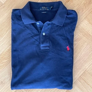 Mörkblå pikétröja från Ralph Lauren - Säljer en stilren mörkblå pikétröja från Ralph Lauren med klassisk krage och knappar. Tröjan har en röd broderad logotyp på bröstet och är i Classic Fit. Perfekt för en avslappnad men ändå snygg look. 